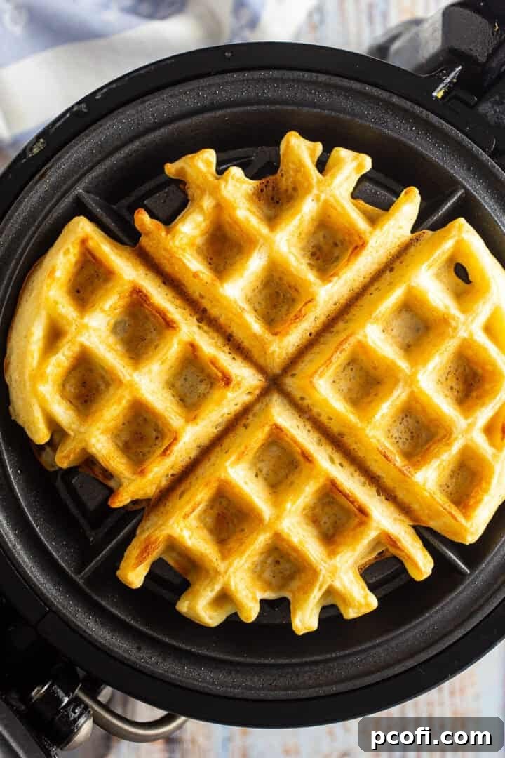 A perfectly golden brown Belgian waffle cooking on a waffle maker, steam gently rising, indicating it's nearly done.