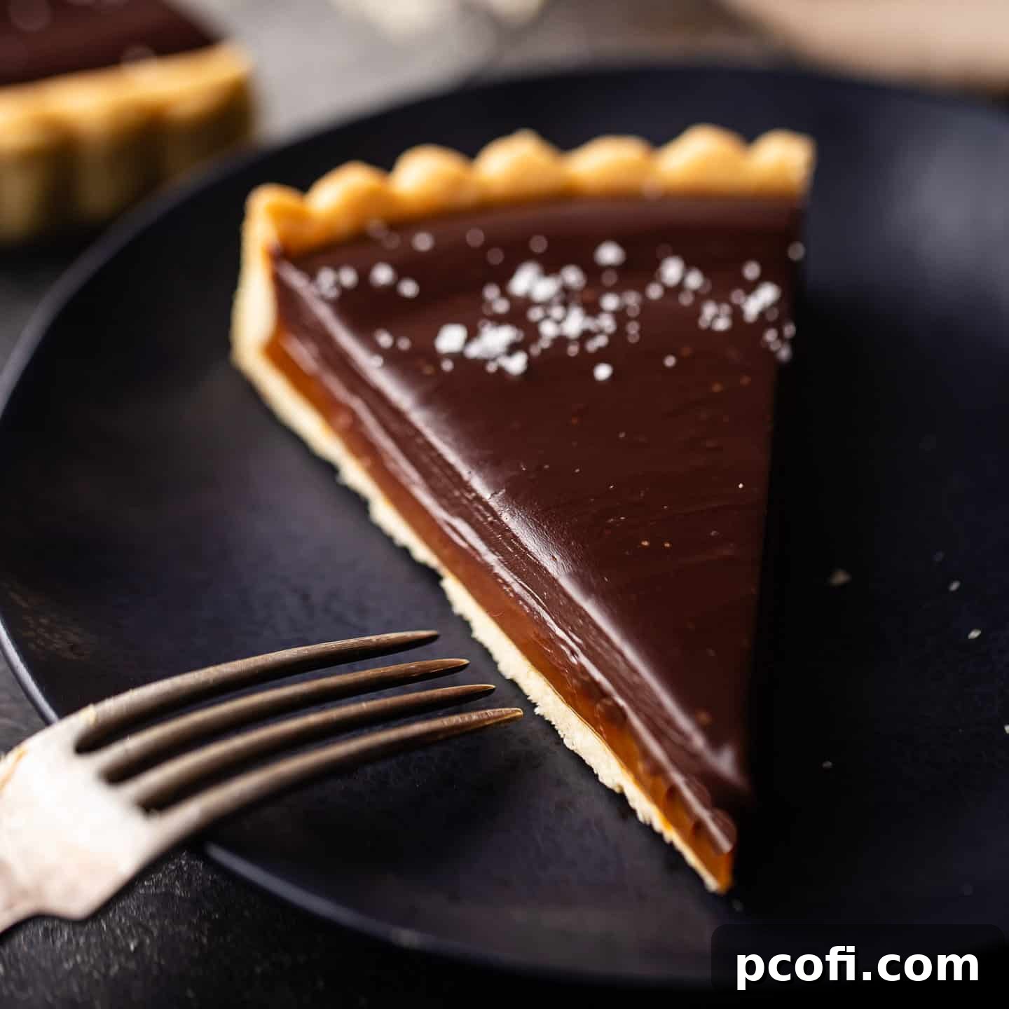 A stunning slice of salted caramel chocolate tart on a dark blue plate, accompanied by a vintage silver fork, highlighting its elegant layers.