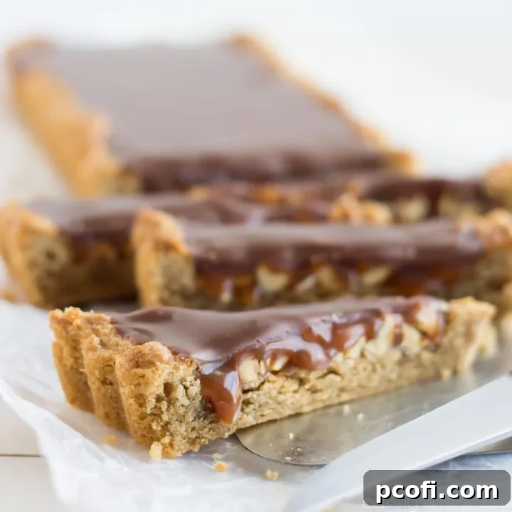 A stunning Snickers Blondie Tart featuring a rich peanut butter blondie base, layered with crunchy peanuts, luscious salted caramel, and a smooth milk chocolate ganache, perfect for a show-stopping dessert.