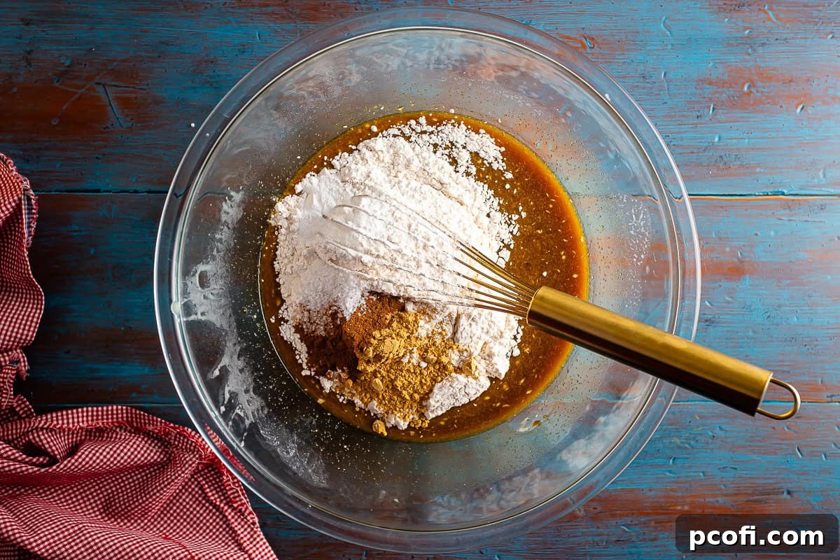 All the dry ingredients for gingerbread cake, including flour, ginger, cinnamon, salt, baking powder, and baking soda, are added into a mixing bowl containing the wet ingredients, poised for thorough mixing.