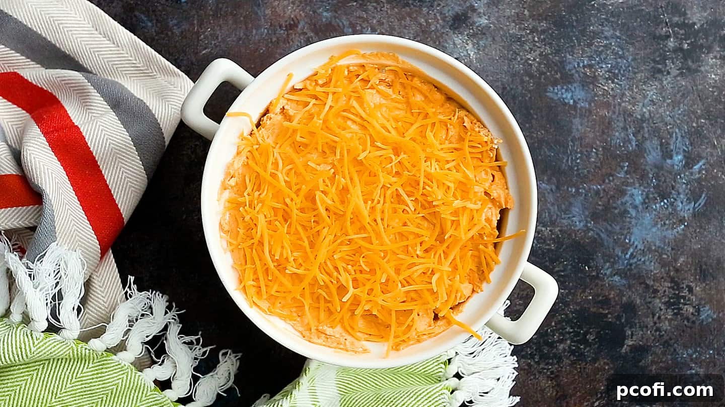 Buffalo chicken dip in a baking dish topped with shredded cheddar cheese, ready for the oven.