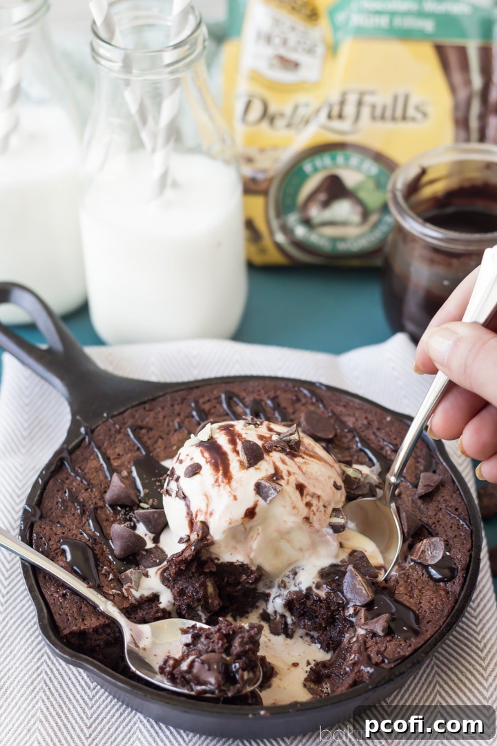 A warm chocolate mint skillet brownie topped with a generous scoop of vanilla ice cream and drizzled with hot fudge sauce. Steam rises from the brownie, highlighting the perfect contrast of temperatures and textures.