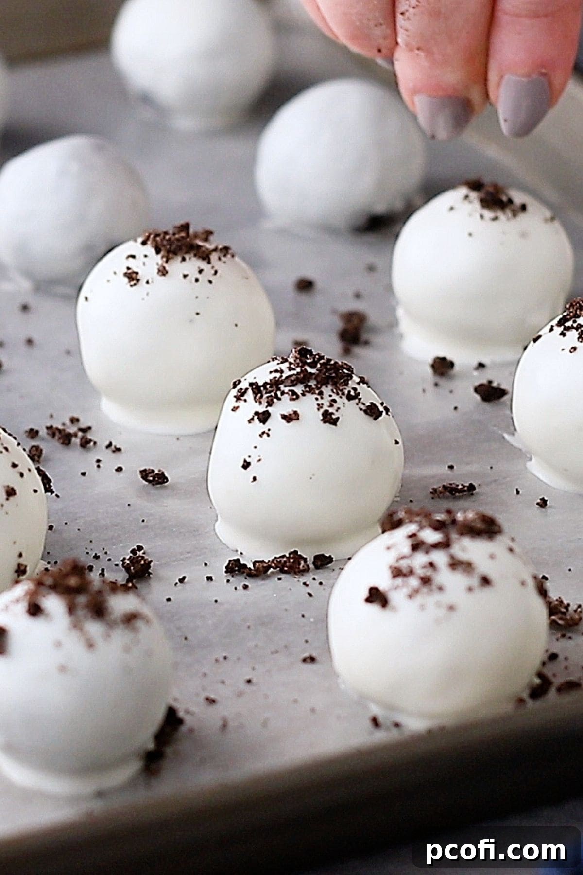 Freshly coated Oreo balls lined up on a baking sheet, being sprinkled with additional Oreo crumbs while the chocolate is still wet for decoration.