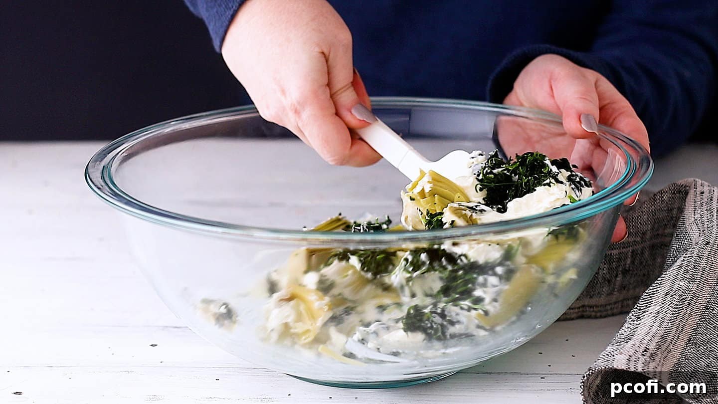Gently folding the spinach and artichoke hearts into the creamy, softened cream cheese base.