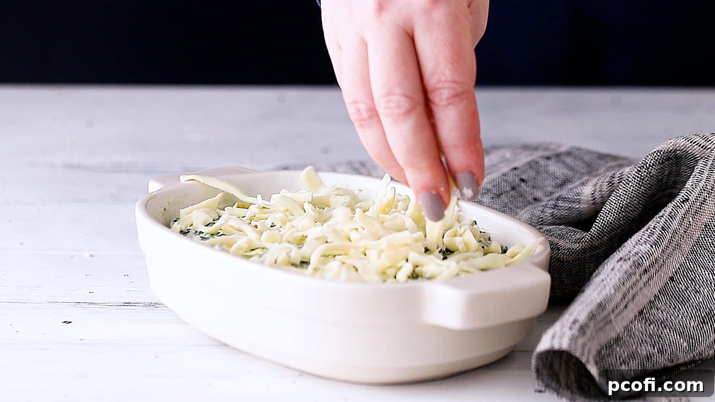 Generously topping the spinach artichoke dip with shredded mozzarella cheese before baking.