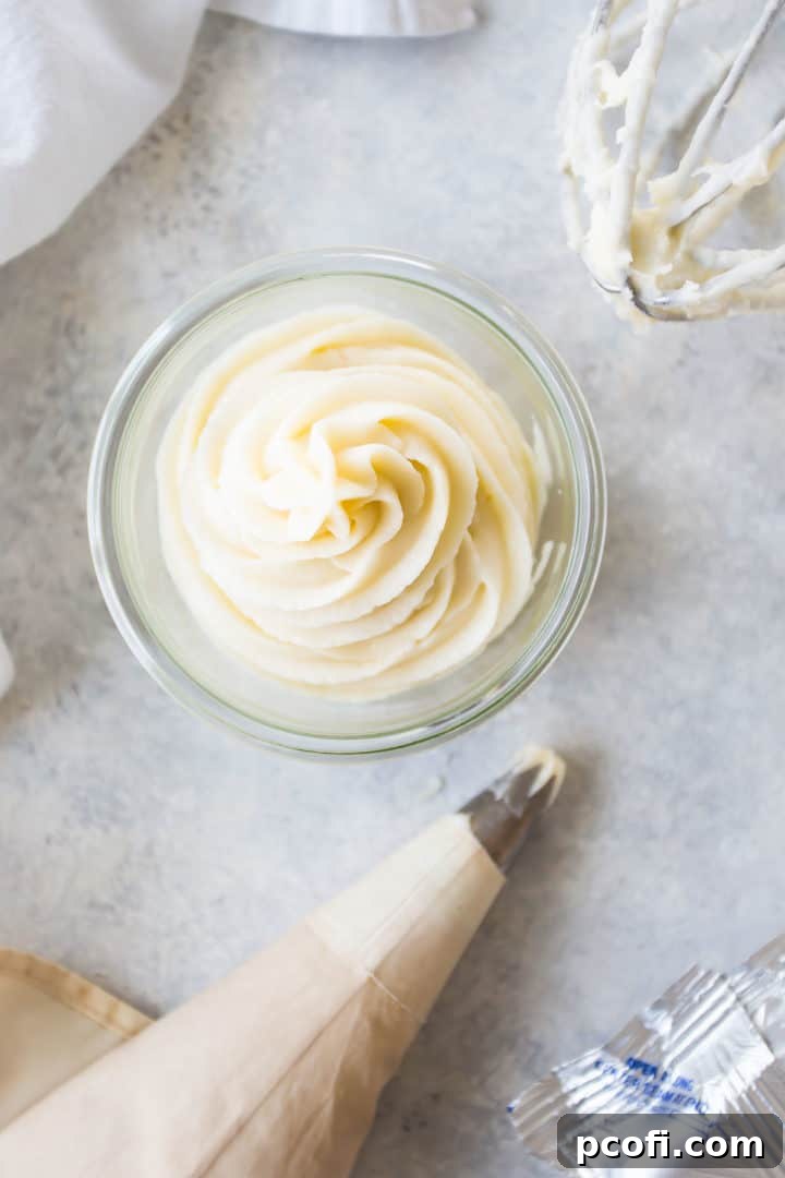 Overhead shot of a bowl of cream cheese icing, a piping bag, and a whisk, with a block of cream cheese in one corner and a white kitchen towel.