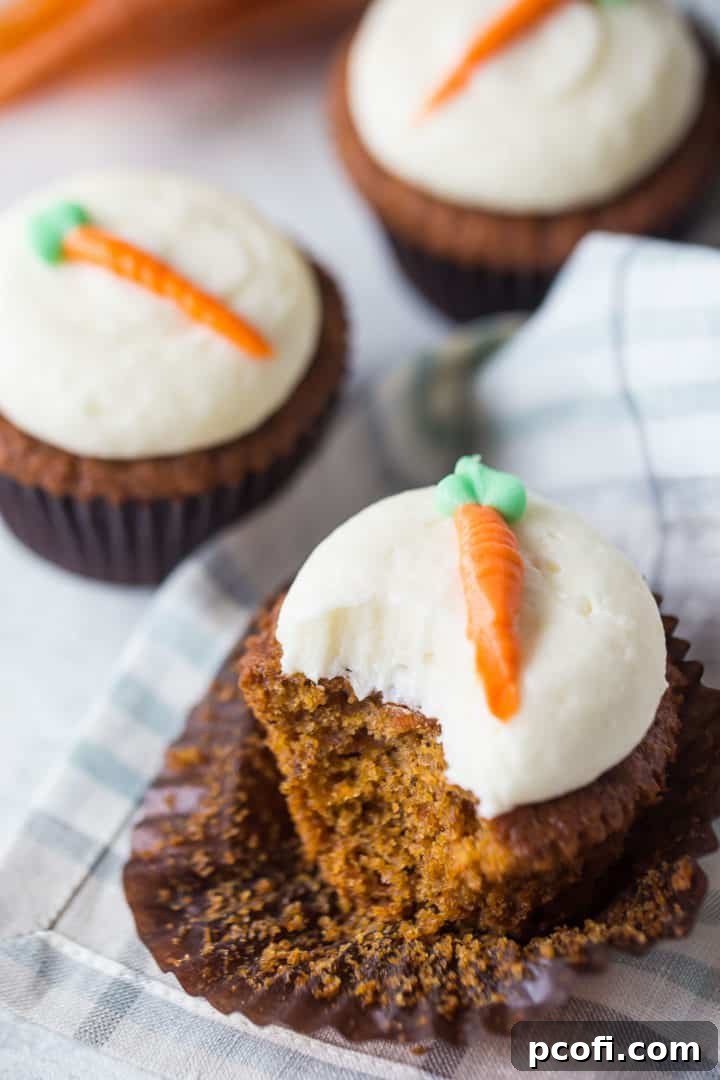 A close-up shot of a carrot cake cupcake with a bite taken out, revealing the moist, spiced interior studded with carrot and pineapple pieces, crowned with a thick layer of cream cheese frosting.