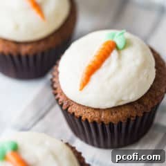 A carrot cake cupcake on a plaid cloth, decorated with cream cheese frosting and an orange carrot piped on top with a green leaf.