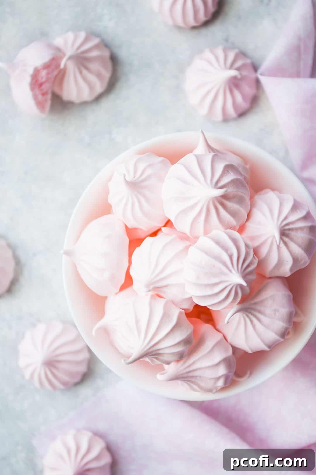 Overhead image of baby pink meringue cookies in a white bowl with a pink cloth, showcasing their vibrant color.