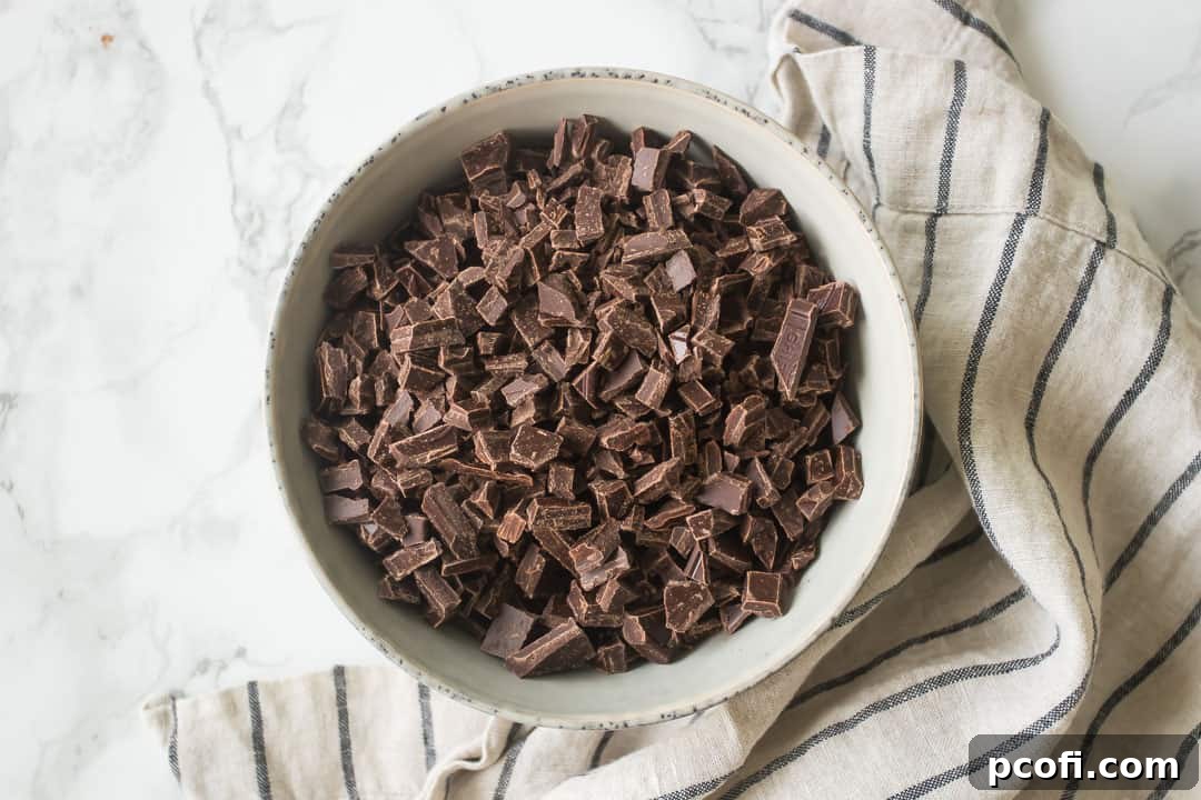 Chopped chocolate in a bowl, awaiting the hot cream, next to a striped kitchen towel.