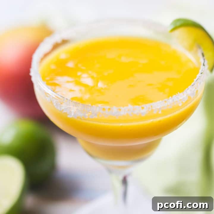 Frozen mango margarita in a salt-rimmed glass with fresh mangoes and limes in the background, signaling a refreshing summer drink.