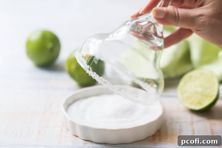 The rim of a margarita glass being gently pressed into a shallow dish filled with coarse kosher salt, creating a perfectly salted rim.