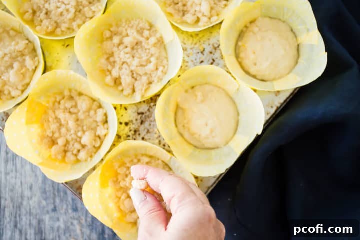 Crumb topping being liberally sprinkled over the unbaked lemon muffin batter in a muffin pan, creating a generous layer of streusel before baking.
