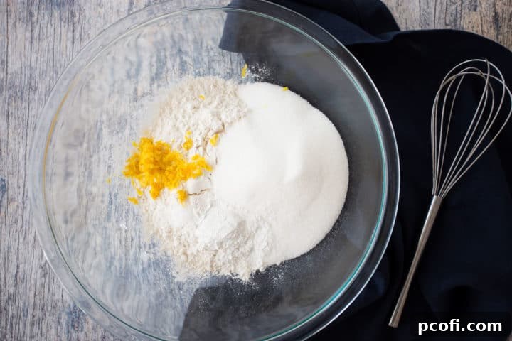 A clear glass bowl filled with the dry ingredients for lemon muffins, including flour, sugar, baking powder, salt, and lemon zest, accompanied by a whisk and a dark blue linen napkin.