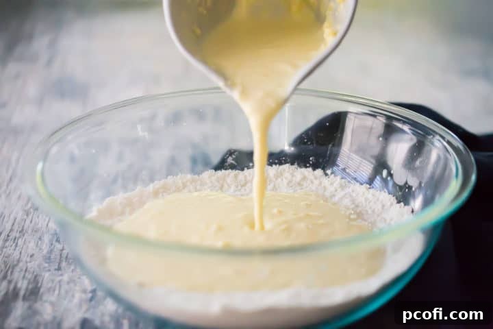 The moment of combining wet and dry ingredients: liquid mixture being poured from a ceramic pitcher into a glass bowl containing dry lemon muffin ingredients, ready for gentle folding.
