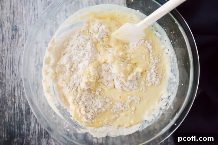 Lemon muffin batter being gently folded with a spatula in a large glass mixing bowl, showcasing its lumpy but well-combined texture, indicative of proper mixing.