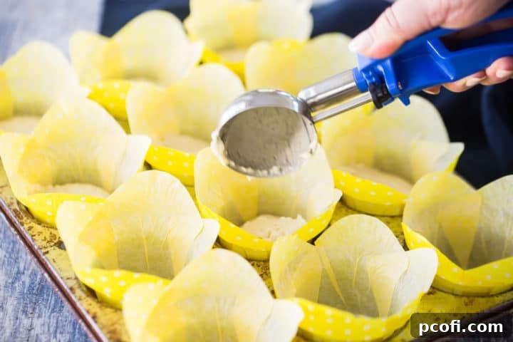 A baker using a 1/4-cup ice cream scoop to carefully portion lemon muffin batter into a muffin pan lined with yellow paper cups, ensuring even distribution.
