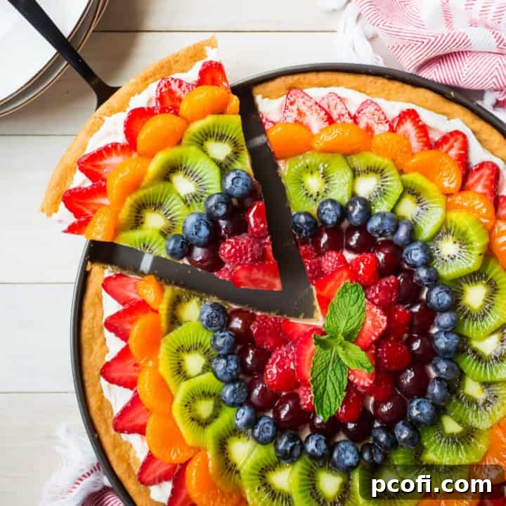 Removing a slice from a rainbow fruit pizza. A vibrant, colorful dessert with fresh fruit, cream cheese frosting, and a soft sugar cookie crust.