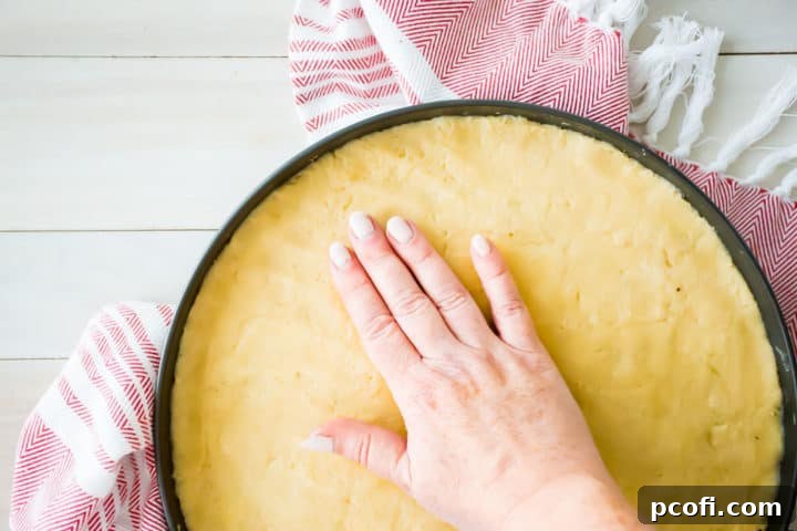 Pressing sugar cookie dough into a pizza pan. Hands gently spreading a golden-brown sugar cookie base into an even layer in a round pizza pan, preparing it for baking.
