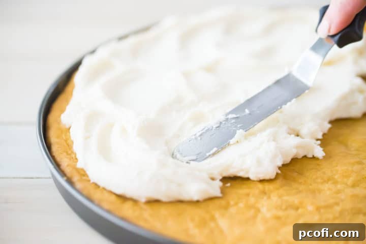 Spreading fluffy cream cheese frosting over sugar cookie crust for fruit pizza. A spatula carefully applies a smooth, white layer of frosting onto a baked sugar cookie crust.
