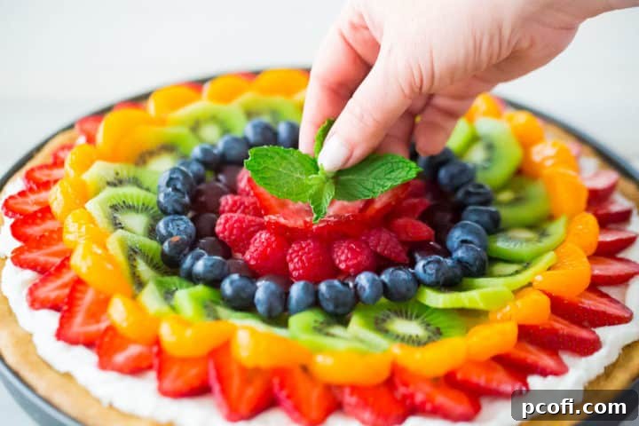 Placing a sprig of fresh mint in the center of a homemade fruit pizza. A hand carefully adds a vibrant green mint sprig to the middle of a colorful fruit pizza, enhancing its fresh aesthetic.