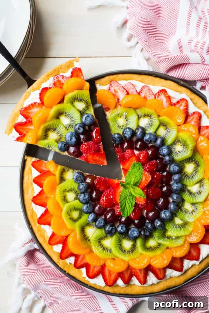 Overhead image of a rainbow fruit pizza with a slice being removed, on a white background with a red printed kitchen towel. A hand lifts a slice from the vibrant, freshly prepared fruit pizza, revealing its soft sugar cookie crust and creamy frosting.