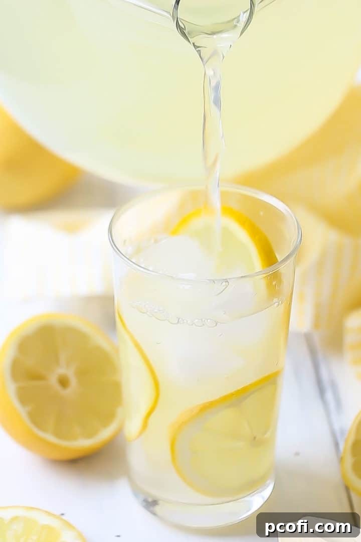 Pouring lemonade from a glass pitcher into a tall glass of ice with lemon slices.