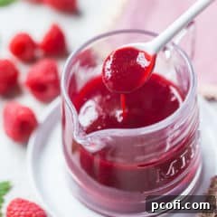 Square image of a small glass pitcher of thick raspberry sauce, being spooned out with a white ceramic spoon.