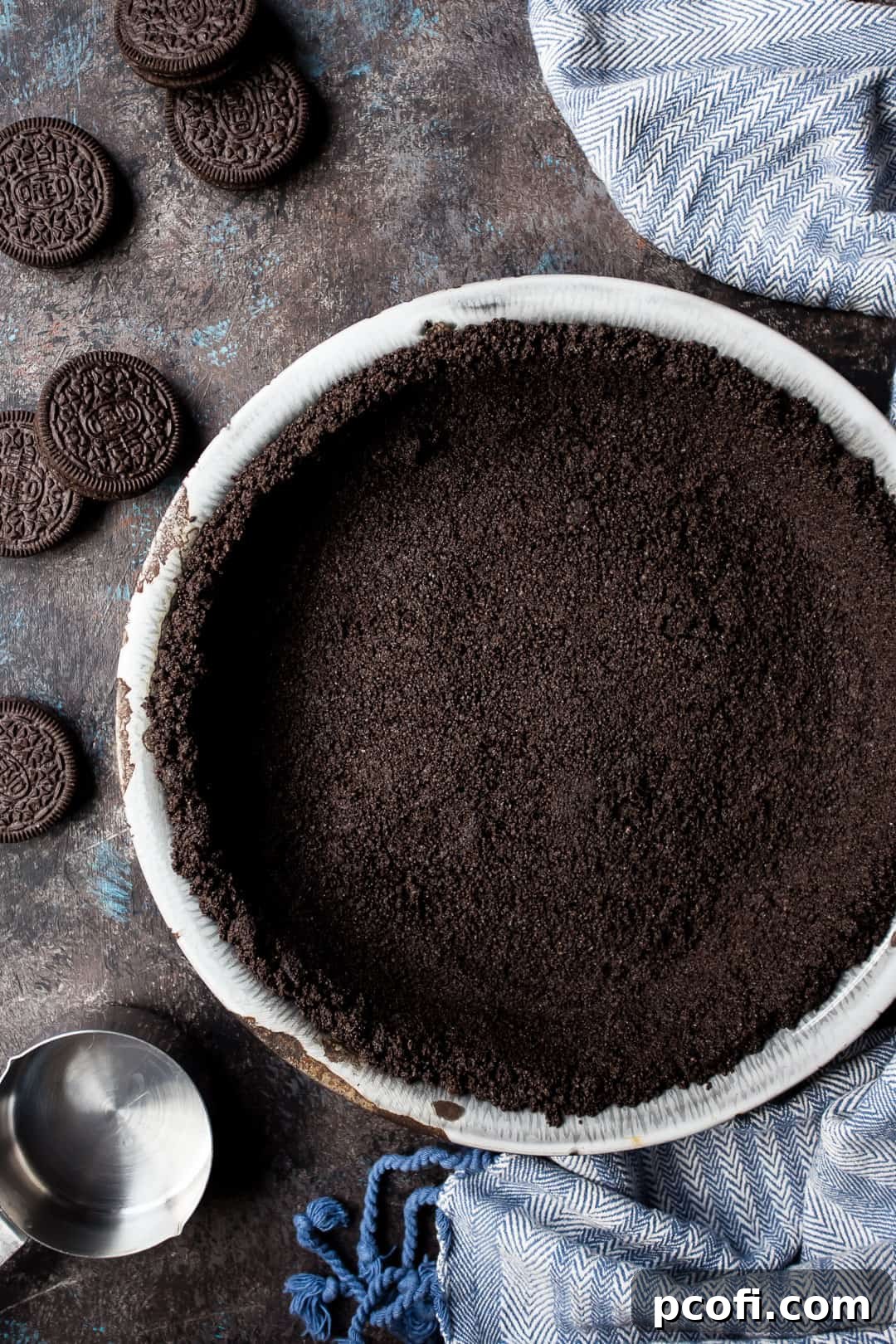 Overhead image of a perfectly formed Oreo cookie crust in a pie dish, surrounded by whole Oreo cookies and a blue cloth, ready for a delicious filling.