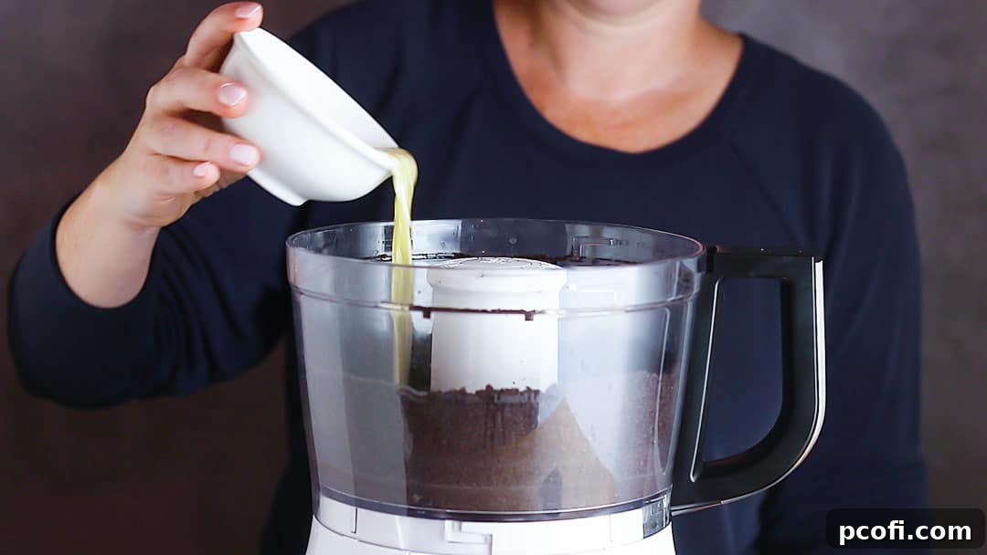 Pouring melted butter over finely ground Oreo cookie crumbs in a food processor, illustrating the next step in making the crust.