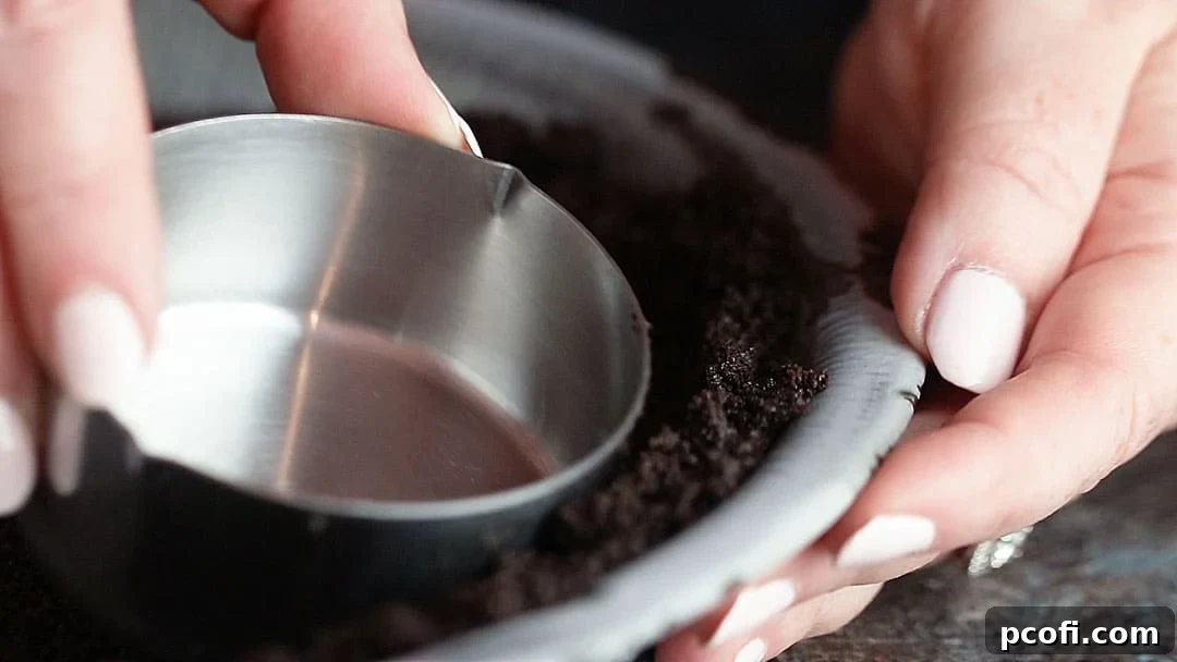 Close up image of shaping Oreo pie crust along the sides of a dish using a measuring cup, ensuring even thickness.