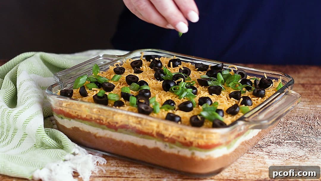 The final step of garnishing a 7-Layer Mexican Dip with sliced black olives and fresh green onions, enhancing its visual appeal and flavor.