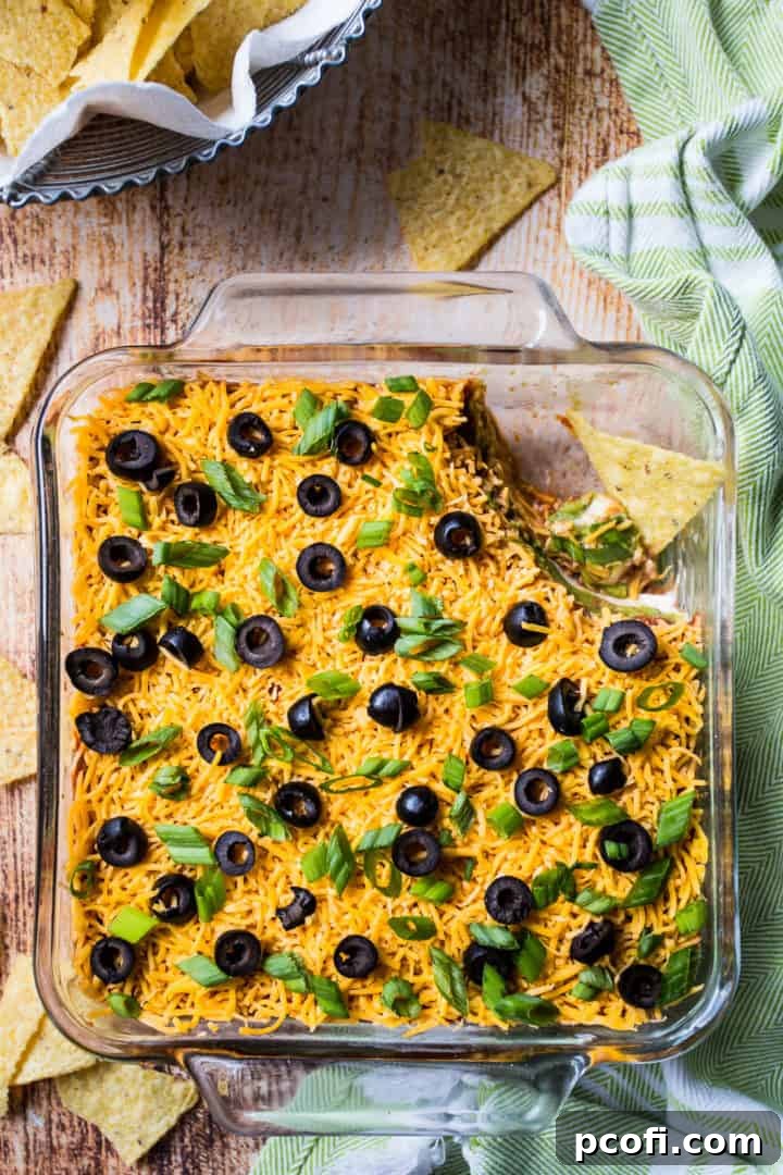 An overhead shot of a perfectly layered 7-Layer Dip, accompanied by a woven basket of crispy corn chips and a vibrant green kitchen towel, ready for serving.