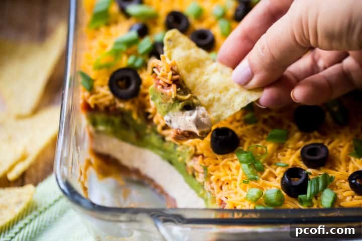 A corn chip perfectly scooping a portion of rich 7-Layer Taco Dip, showcasing its creamy texture and distinct layers.