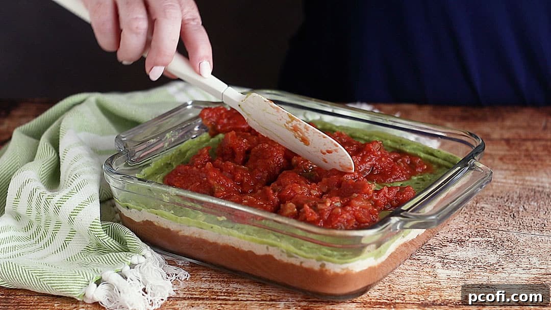 Applying a layer of strained salsa evenly over the guacamole, ensuring a vibrant and flavorful addition to the dip.