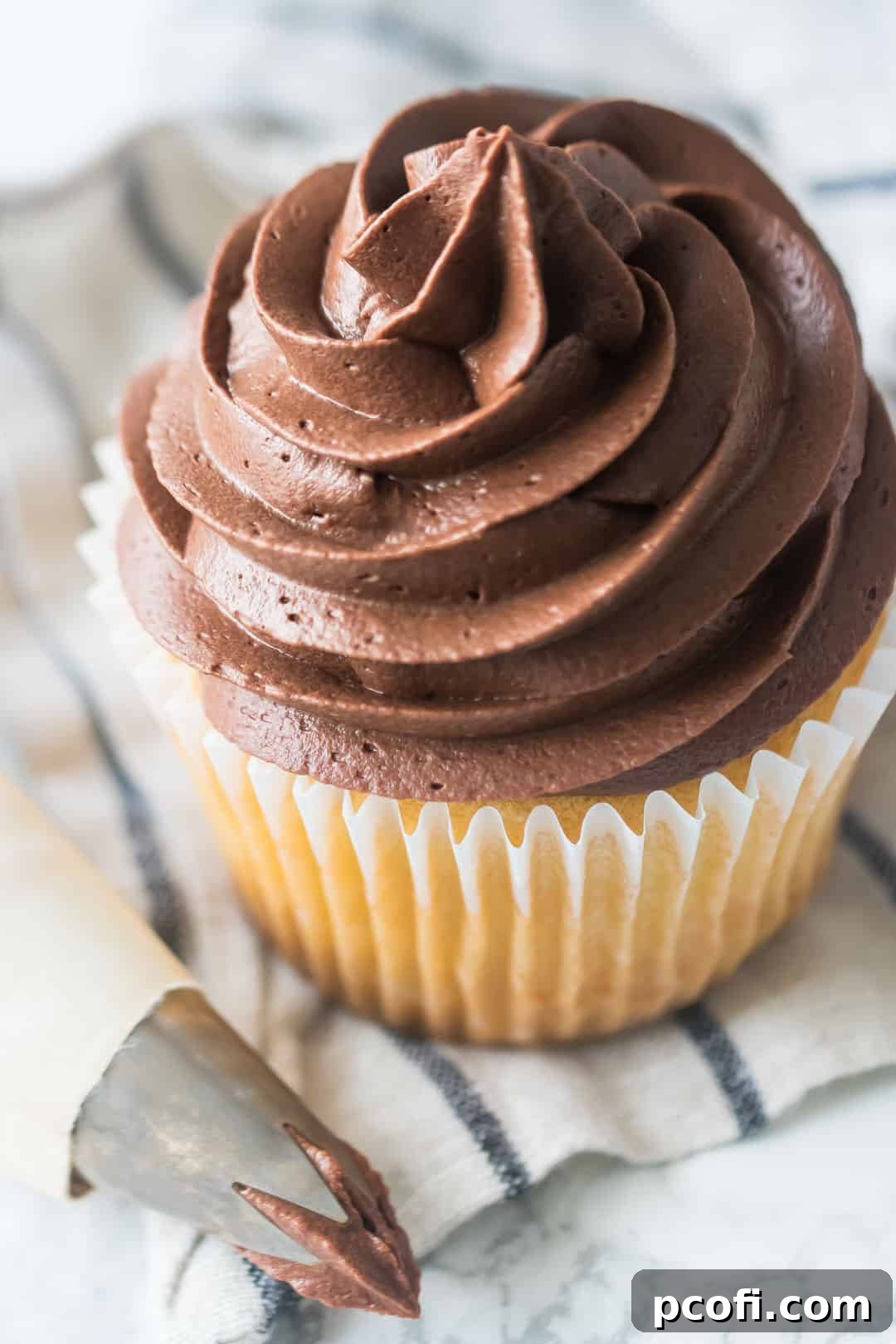 A yellow cupcake adorned with luscious whipped chocolate ganache frosting, resting on a striped fabric, with a piping bag nearby.