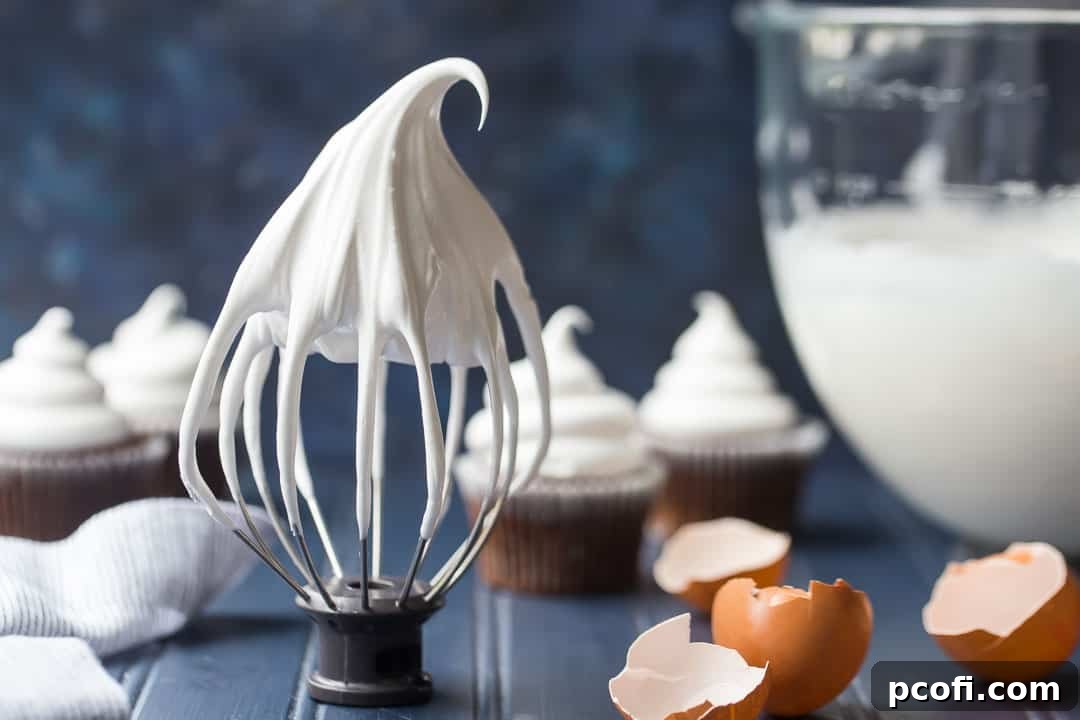 A whisk attachment covered in fluffy marshmallow frosting, with blurred eggshells and cupcakes in the background, hinting at the baking process.