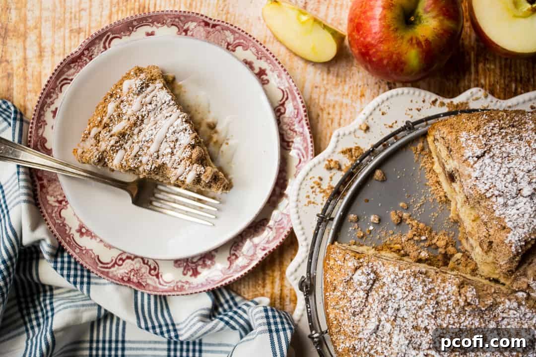 An inviting overhead view of a single, perfectly sliced piece of apple sour cream coffee cake, with the remainder of the whole cake artfully positioned to one side.