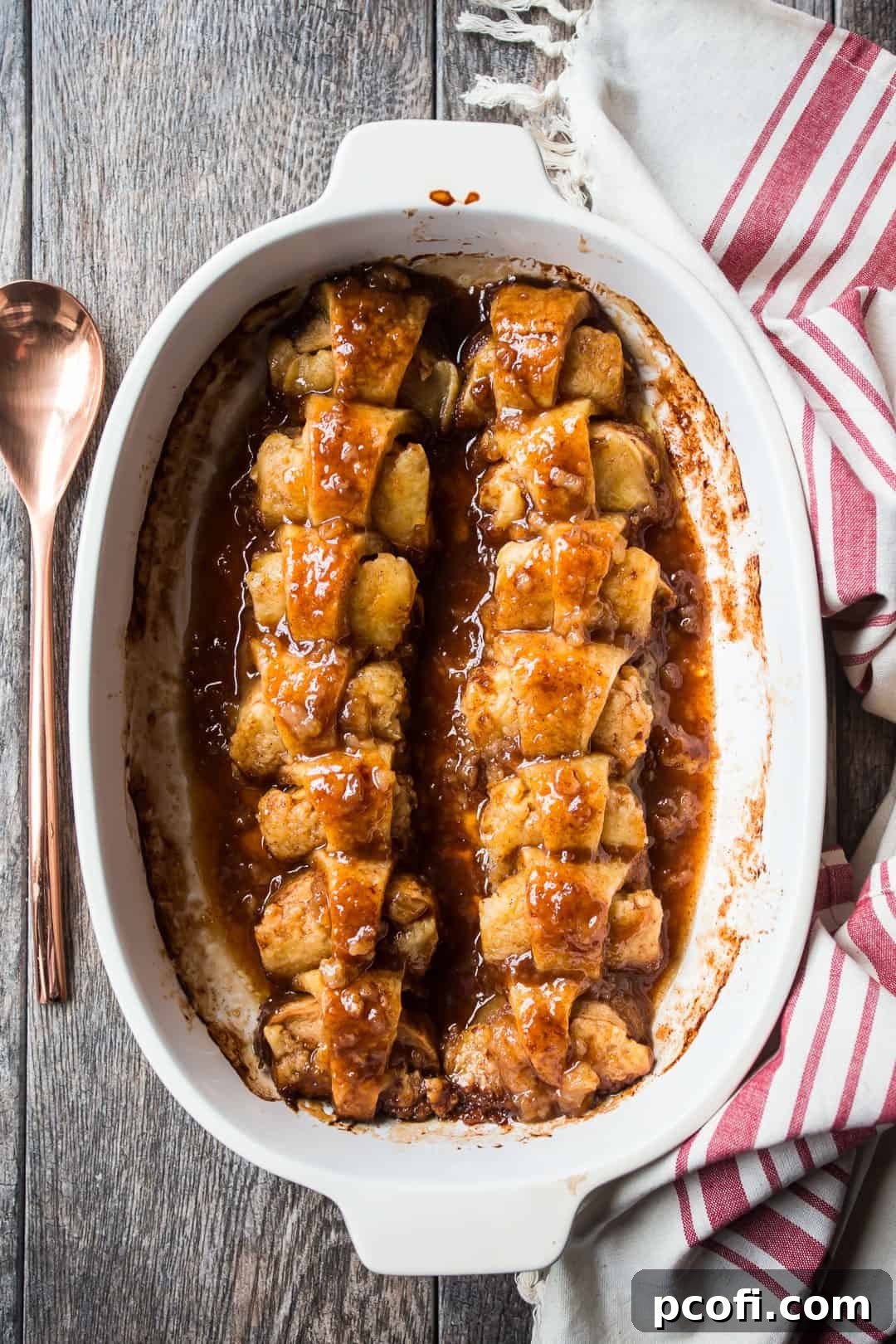 Crescent roll apple dumplings in a ceramic baking dish with a copper serving spoon and a linen kitchen towel. A warm, baked dessert.