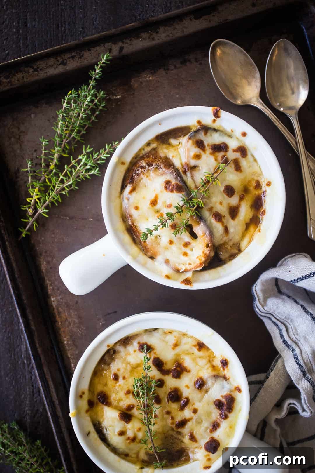 Overhead image of two bowls of French onion soup on a brown tray with a striped cloth, vintage silver spoons, and a sprig of fresh thyme.