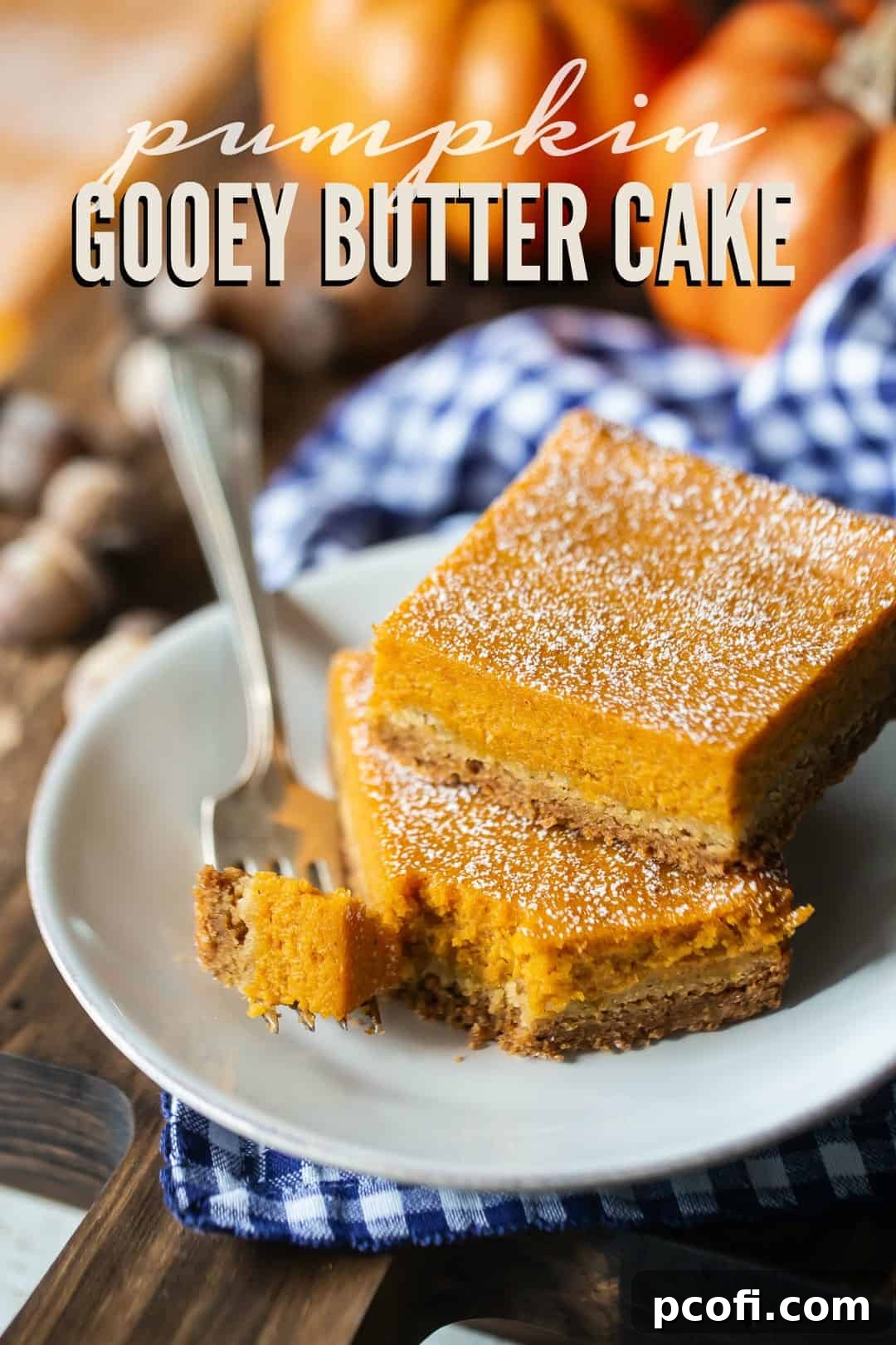 Close-up of golden-brown pumpkin gooey butter cake bars on a white plate, garnished with a blue checked napkin and a text overlay above that reads "Pumpkin Gooey Butter Cake," highlighting its homemade appeal.