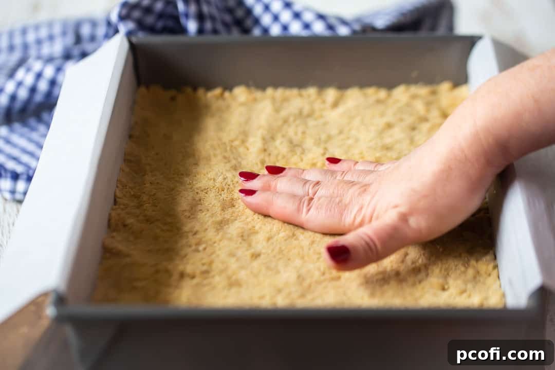 Hands pressing the crumbly bottom layer mixture evenly into a greased and parchment-lined 9x13-inch pan, preparing for the pumpkin gooey butter cake filling.