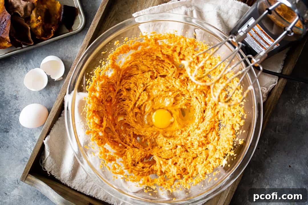 Cracked eggs being carefully added to the vibrant sweet potato pie filling in a glass bowl, ready to be blended into the mixture.