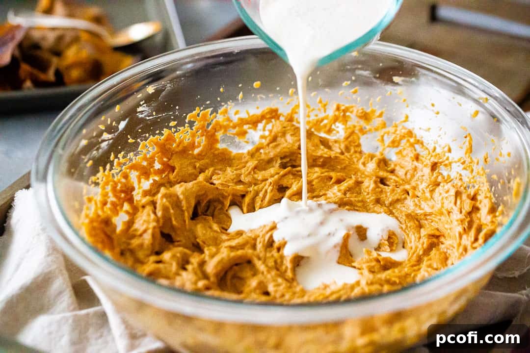 A stream of rich heavy cream being poured into the spiced sweet potato filling mixture in a large glass bowl, adding to its luxurious texture.