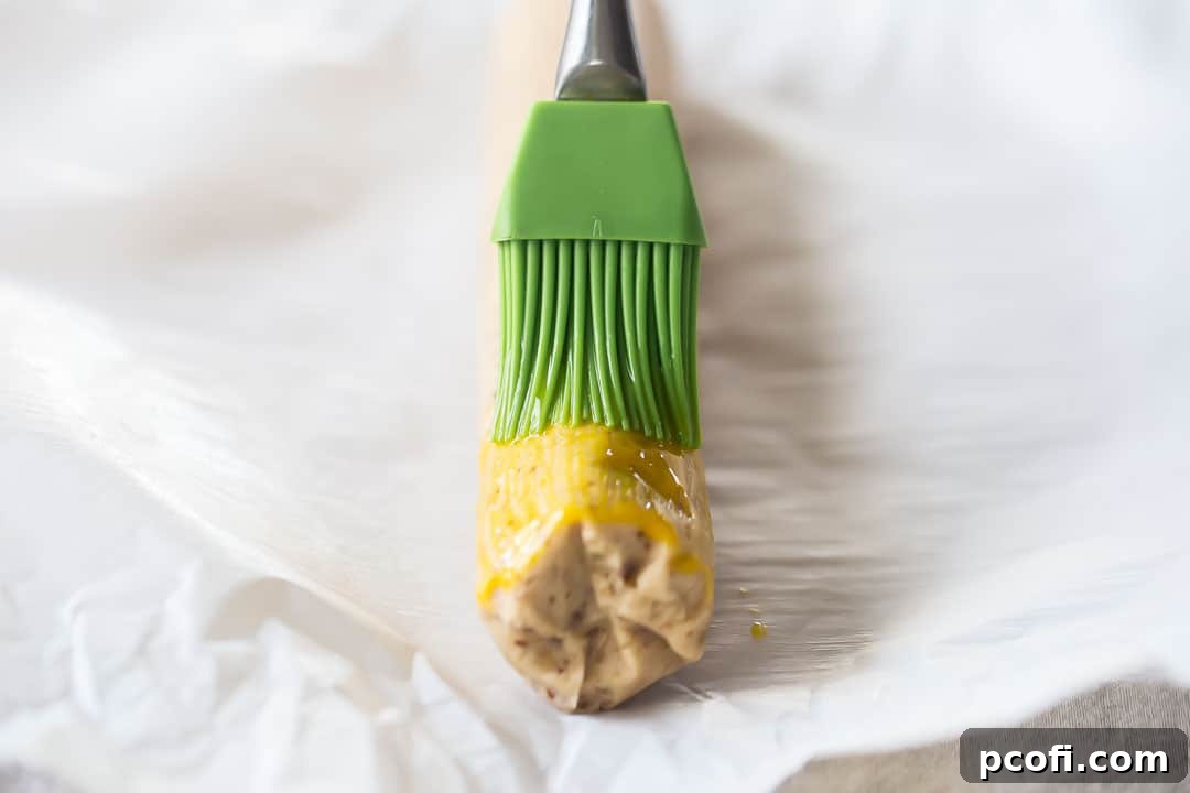 A chilled pecan sandies cookie dough log being brushed with the remaining egg yolk, preparing it for the pecan coating.