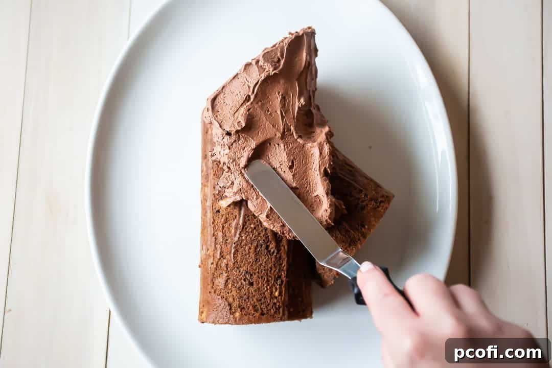 Applying a thick, textured layer of whipped chocolate ganache frosting onto a yule log cake using an offset spatula, creating a bark-like finish.