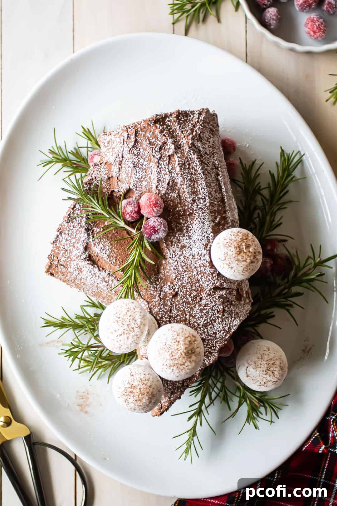 Overhead shot of a beautifully decorated chocolate yule log cake (Buche de Noel) adorned with powdered sugar, delicate meringue mushrooms, fresh green rosemary sprigs, and vibrant sugared cranberries.