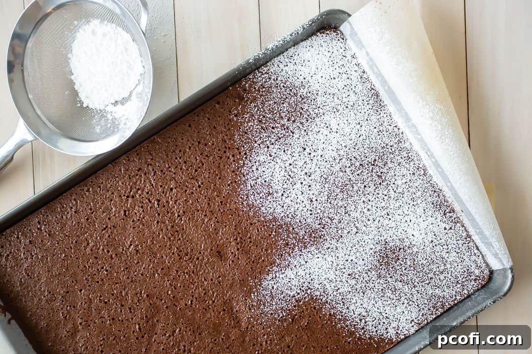 Generously dusting a warm chocolate yule log cake with powdered sugar immediately after baking, a crucial step for easy rolling.