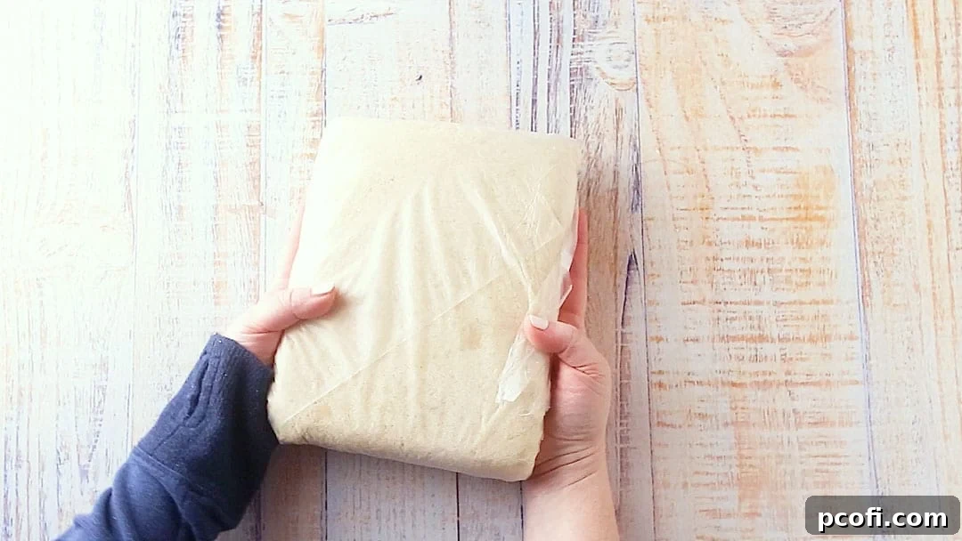 Shortbread cookie dough compactly pressed into a flat square and tightly wrapped in plastic wrap, ready for chilling.