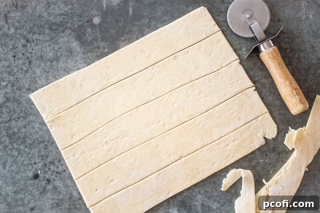 A hand using a knife to cut long, even strips of puff pastry dough.